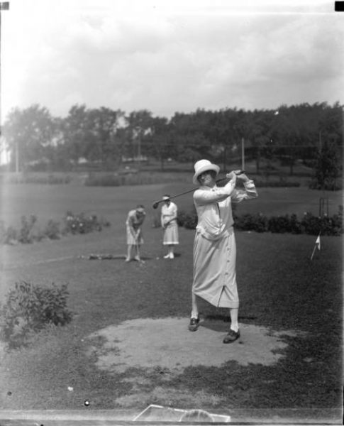 Grayscale long shot of a woman showing a golf swing movement on a Milwaukee course. Two women play several feet behind. They all wear casual clothes and hats.