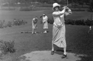 Grayscale long shot of a woman showing a golf swing movement on a Milwaukee course. Two women play several feet behind. They all wear casual clothes and hats.