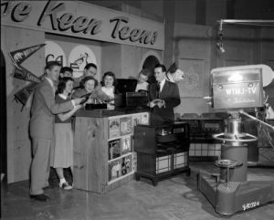 Grayscale full shot of eight Milwaukee high school students smiling as they pose with vinyl records in WTJM-TV broadcast studio. They stand on the left while facing to the right behind a high table with record players atop. Behind them is the studio set with the name "The Keen Teens" inscribed on it. A vintage camera with the WTJM-TV logo and an overhead mic are visible on the image's right.