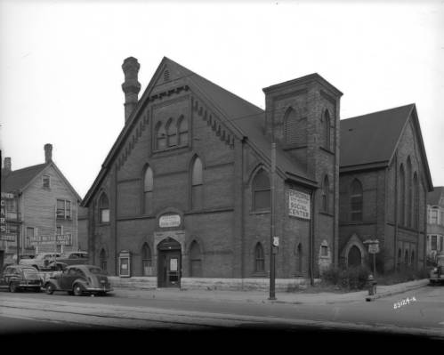 Sepia-colored long shot of Episcopal Social Center's facade facing slightly to left on a street corner. The front section, which consists of two-and-a-half stories, features an entrance and multiple three-pointed arched windows. The Center's name signage hangs on top of the front door and on the exterior wall facing the other roadside. Cars are parked in the left foreground, next to the building.