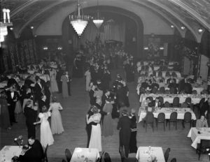 High-angle shot of a group of pairs dancing in Chrysanthemum Ball. The couples stand in suits and gowns in a dance position on the floor. An array of dining tables flanks them. The lights are on under the vaulted ceiling.