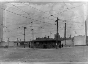 Long shot of a single-story streetcar station on a street corner. Some people stand next to the building, some walk pass the station. Above are the crisscrossing overhead wires. The streetcar tracks embedded in the street are in the foreground.