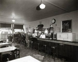 Grayscale long shot of a tavern's interior with tables and chairs in the left foreground, the bar and stools in the background. A group of people sitting on stools and two people standing behind the bar make eye contact with the camera. Shelves with bottles of liquor are visible in the back. A clock on the wall reads 10:40 and daylight is visible through the window.