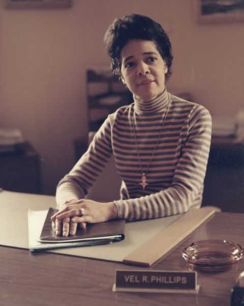 Medium shot of Vel Phillips in a striped sweater and a necklace sitting behind a desk that is placed in the image's foreground. Her body faces the camera lens while her eyes glance slightly to the right. Phillips lays her hands atop documents on the desk. A nameplate with her name is in the center foreground. On its right behind is an empty glass ashtray. The background is blurred, showing a wall and some office supplies.