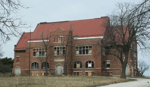 Long shot of the former Milwaukee County School of Agriculture and Domestic Economy building. The multiple-story abandoned structure has its windows and doors covered. A barren wide yard and leafless trees are set in front of the building.