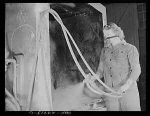 Grayscale medium shot of a Heil Company employee spraying small parts for gasoline tanker trucks in the left. The person in a mask and uniform stands on the right.