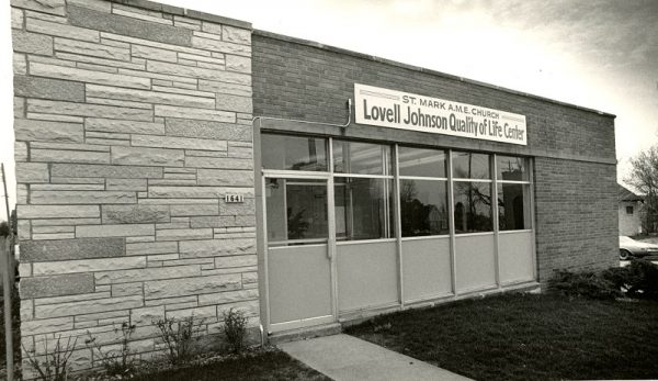 A grayscale long shot of the facade of St. Mark A.M.E. Church Lovell Johnson Quality of Life Center. The image shows the entrance of a one-story building with a closed door and windows. A small address sign on the wall displays the number "1641."
