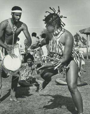 Image of a drummer and a dancer performing in an outdoor space. On the left is the shirtless man with a drum strap on his neck and a djembe resting between his legs. His palms float in the air ready to hit the drum. On the right side is the female dancer moving her body expressively in an animal print costume. Her braided hairs fly in the air; her hands and one of her legs are floating in a dance motion.