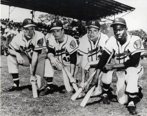 Sepia-colored photograph of four members of the 1957 Milwaukee Braves kneeling on a baseball field while holding bats. They pose in uniforms. Blurred in the far background is a grandstand full of spectators.