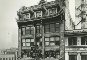 Facade of the Toy Building by a sidewalk in grayscale. The image showcases its six-story structure with a touch of Chinese roof architecture. Chinese letters adorn its exterior walls. The building features a long canopy on the ground floor and a balcony on the second floor. A sign reading "Toy Chop Suey Dancing" projects from the building's third story.