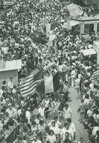 Grayscale high-angle shot of the 1980 Festa Italiana procession winding through a huge crowd of people. Some participants carry Italian and American flags, religious statues, and banners.