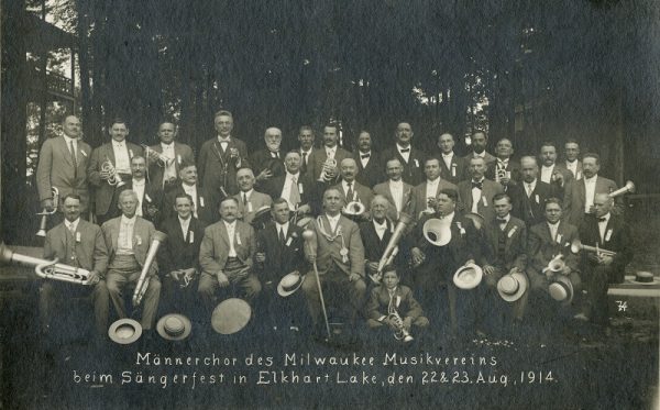 Three rows of the men's chorus of the Milwaukee Musikverein pose in a sepia-colored photo. Men in the two front rows sit, and the last row stands, all in their suits and ties. Most of them hold a brass instrument. A man sitting in the middle front carries a baton.