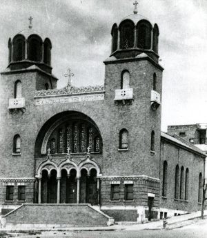 Exterior view of the original Greek Orthodox Church of the Annunciation in grayscale. The facade features a grand entrance that has portals adorned with arched structures. Two gate towers flank the front doors. Crosses stand atop the towers. Words in Greek are inscribed along the center roofline.