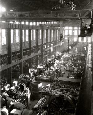 High-angle shot of the interior of the Allis-Chalmers' factory building. Rays of sunshine from large windows surrounding the walls light up different gears and machinery equipment lying on the floor. Some machine parts are stacked on top of each other, and some are kept in boxes. When zooming the image, men standing and working beside the huge machine parts come into focus. Above them is a crane hook hanging on the tall ceiling. Close to it, at the top right corner of this image, is a man standing with one hand on his hips and the other hand holding a glass. He seems to oversee the workers below.