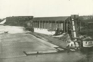 A drawing illustrating a small group of people cutting a large sheet of ice atop a body of water into smaller blocks. Some blocks are loaded onto an inclined conveyor belt on the right. A building with the sign "Wis. Lakes Ice & Ctge. Co." is visible in the background.