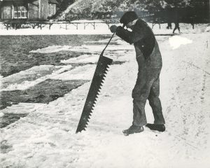 20th century photograph of a man cutting chunks of ice from a river with a large saw. 