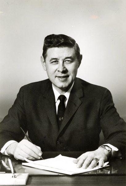 Medium shot of Henry W. Maier smiling in a formal attire while sitting behind a table facing directionally to the camera lens. He poses in a writing position with right hand holding a pen and left hand on top of a document.