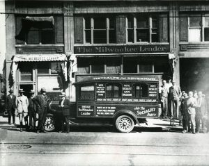 Long shot of a group of people standing outside the Milwaukee Leader facade in sepia. Most of them pose in formal attire, flanking the company's delivery truck covered with messages promoting the Socialist Party. The image shows the office's ground and second floor.