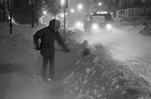 Grayscale full-shot of someone in a winter coat shovelling the sidewalk. Snow piles appear along the sidewalk in the foreground to the background. Snows cover the street that stretches down on the far right. A bus with lights on runs on the street. The streetlights glow in the background.
