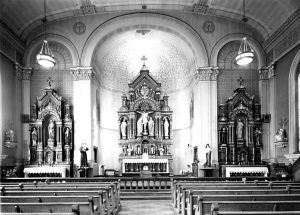 Grayscale long shot of three altars at the Holy Trinity Our Lady of Guadalupe church. Each altar stands in front of an arched structure on the wall. The central one has the largest structure. Rows of pews appear in the left and right foreground, flanking an aisle. Two ceiling lights glow on the left and right sides under the vaulted ceiling.