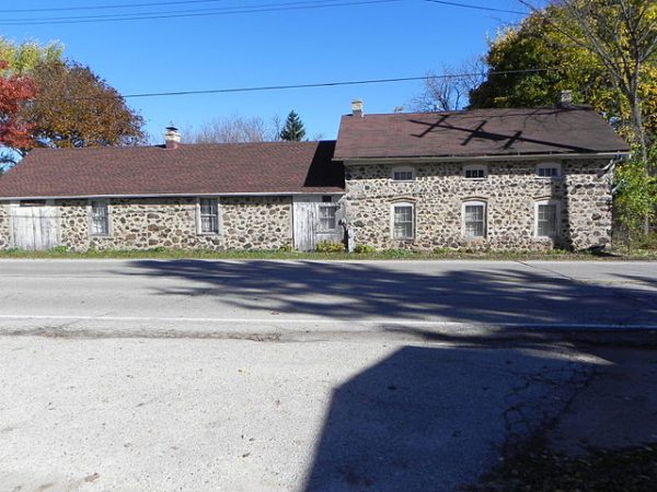 Long shot of the facade of Ritger Wagonmaking and Blacksmith Shop. The building consists of two wings, one that is a single story and one that is two stories with regularly spaced windows. It has a brown roof and walls made of field stones, set near trees beginning to turn with fall colors.