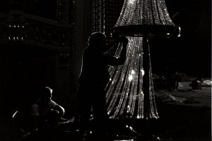 Silhouette of two people working on a large crystal chandelier in the Pabst Theater. The image is mostly pitch black. One of the people stands at the image's center with hands touching the sparkling crystals. Arrays of various smaller lights form two large arches in the background.