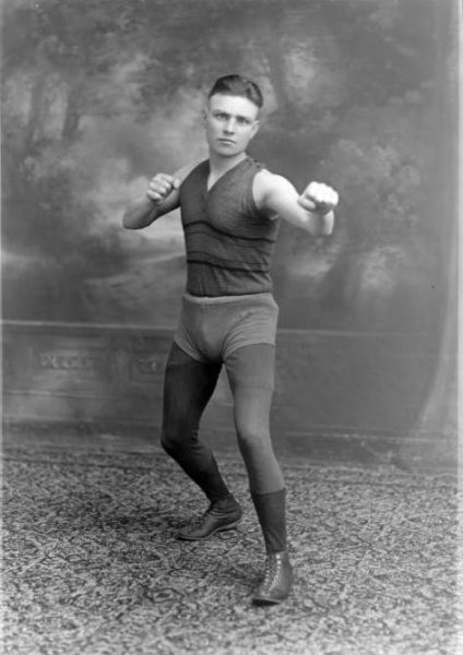 Grayscale full shot of Anton Chmurski demonstrating a boxing motion with eyes staring directly into the lens. For this studio portrait, he wears a sleeveless shirt, short pants on top of tight-fitting pants, socks, and shoes.