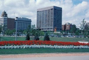 The large Northwestern Mutual building appears at the image's center in the distance, set between other lower buildings. O'Donnell Park's iconic artwork named "The Calling" and a parking lot filled with cars are visible near the tower. Next to the parking area is the park's green scenery adorned by bright red landscaping plants lining from left to right foreground. A cloudy sky is in the background.