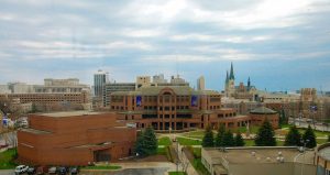Elevated view of a building complex pm the Marquette University campus and its surrounding area. The Alumni Memorial Union is seen at the image center. Green lawns and landscaping plants arranged in different shapes adorn the yards between buildings in the campus area. Milwaukee's buildings appear in the far background under the blue sky. The Milwaukee County Civic Center and Jail are visible in the left background.