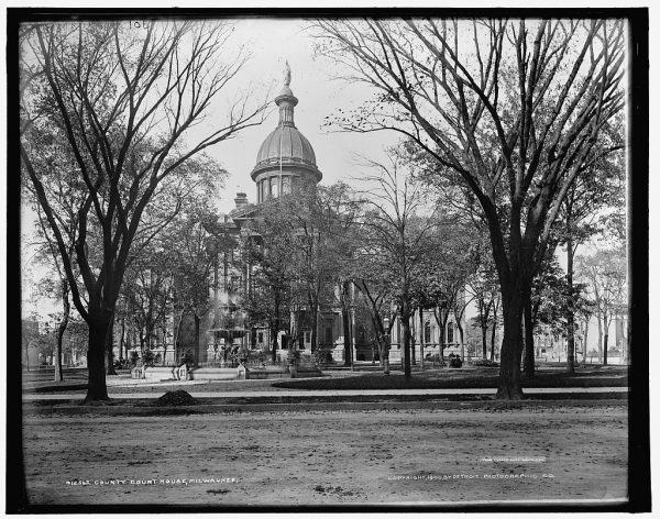 Grayscale long shot of the old Milwaukee County Courthouse's facade hidden behind a line of trees. The building's tower soars above the trees. A water fountain is set in front of the courthouse.