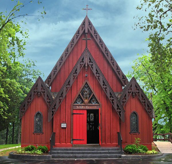 A long shot of the St. John Chrysostom Church facade features the building's board and batten exterior walls in red. The main entrance doors in a brighter red are in the middle; the right door is open, showing stained glass windows in the interior. The church has steep rooflines and crosses installed atop.