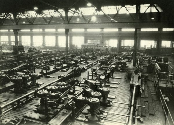 Long shot of A.O. Smith assembly facilities' interior in grayscale tone. Different machines fills up the largely empty building. The ceiling lights are on, glowing like big dots amid the bright shine emanating from the regularly spaced window walls in the background.