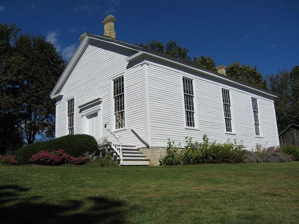 Low-angle shot of the Reformed Presbyterian Church of Vernon facade set on a green lawn. The image shows two sides of the one-story building. The main entrance is on the left, flanked by two rectangular windows. On the right are three windows in the same style. The entrance door and exterior wall are made of wood in white paint. Two stone chimneys are on its roof.