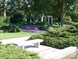 Image of a portion of Boerner Botanical Gardens showcasing its green landscape. Different flowers, bushes, and grass grow on the ground. A group of purple landscaping flowers is set under the shade of a big lush tree in the background. A white gazebo is visible in the distance behind them. A white stone bench stands in the foreground.