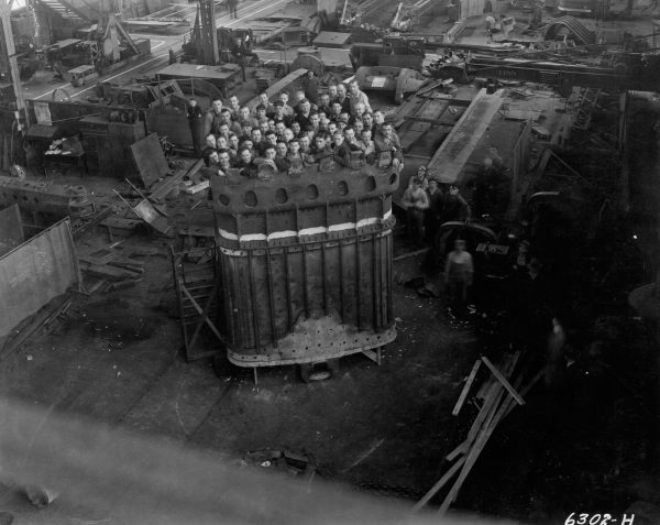 Grayscale high-angle shot of 52 men standing inside a massive dipper for a Bucyrus-Erie 950-B stripping shovel. Appearing in the background are abundant kinds of equipment filling the plant's interior.