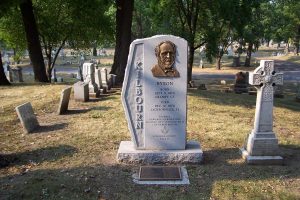 Wide shot of Byron Kilbourn's tombstone in white features a bronze-colored engraving in the shape of his face. His last name is inscribed vertically on the left portion of the stone. Numerous gravestones appear in the background under the shade of trees.