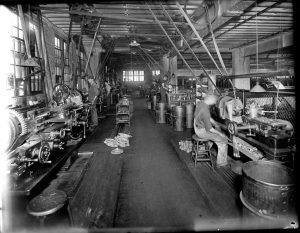Grayscale photo of the Chain Belt Company's interior filled with machinery equipment and gears placed on the left and right sides of the room. An aisle is visible along the center of the room between the machines. Some men sit and some stand while working at metal lathes.