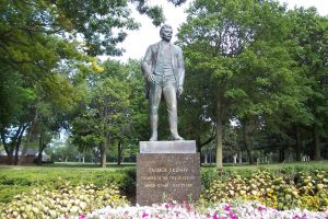 A full shot of Patrick Cudahy's bronze statue, standing and looking to his left. The statue stands in a green landscape. The base reads "Patrick Cudahy, Founder of the City of Cudahy" above his date of birth and death. Colorful landscaping flowers are set below the sculpture. Green lawn and lush trees are visible in the background.