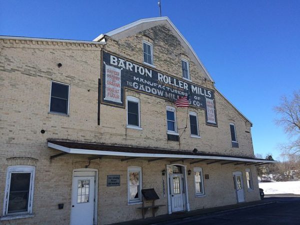 The facade of Barton Roller Mills against the blue sky. The building consists of two and one-half stories and features several white-framed rectangular windows and cream-like coloured exterior walls. Three white doors are placed on the first floor under a wide eave. A small American flag is displayed on the second floor under the mill's huge nameplate.