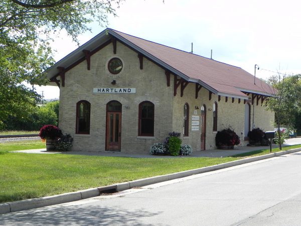 Long shot of the Hartland Railroad Depot. Cream City bricks compose its single-story structure. The image shows two sides of the building. The left side features an entrance, two arched windows, and a bullseye window under the gable roof. The right side also has a door and two windows. A green lawn surrounds the depot that stands by a road.