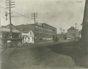 Sepia-colored long shot of the F.C. Gross Brothers Company building on the center back and the R. Gumz & Company on the far right back. Both stand on the left side of a long roadway stretching from right background to foreground.
