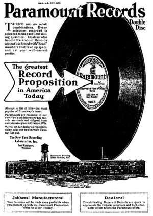 A full page Paramount Records advertisement from the Talking Machine World magazine. The ad is titled with the brand's name. Beneath is a combination of text and a large picture of a black-colored phonograph record with the Paramount logo in the middle. An arrow aiming toward the record contains text that reads, "The greatest Record Proposition in America today." Below is a drawing illustrating the company's plant.