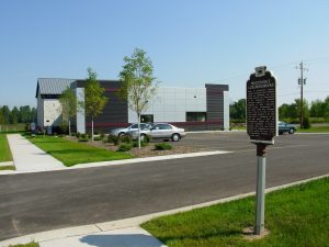 Long shot of a historical landmark sign entitled "Wisconsin's Luxembourgers" containing a short description, facing left. The plaque stands on a green lawn next to an asphalt road stretching in the foreground. The Luxembourg American Cultural Center and a parking lot with three cars are in the background.