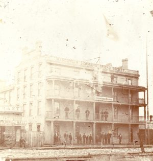 An overexposed sepia-colored photo of the four-story Temperance House building. All upper floors have balconies enclosed with balustrades. Several people stand on the balconies. More appear on the porch. Atop the building is inscribed "Wisconsin Seaman's Friend Society." The Temperance House sign is attached to the third's floor's balusters. Another building appears on the left.