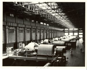 Elevated view of the interior of Lakeside Power Plant's turbine room in grayscale tone. A row of turbine machines appears in the center of the room, from the foreground to the background. Light emanates from the ceiling. The steel structure on the ceiling and the wall lining the left side of the room are visible. Some workers stand on the floor in the right foreground.