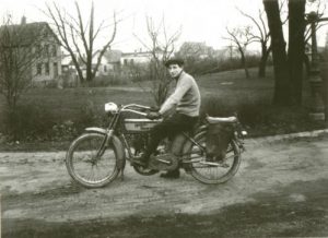 Sepia-colored long shot of a man in a sweater and trousers sitting on a vintage Harley-Davidson motorcycle. He poses with his hands on the handlebars, making direct eye contact with the camera lens. Lawn and leafless trees are in the background.
