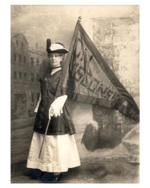 Sepia-colored full shot of Theodora Winton Youmans posing in front of a painted backdrop. In a dress and a hat, Youmans stands facing the left while holding a fanion-shaped banner that reads "On Wisconsin" with her left hand. She makes direct eye contact with the camera lens.