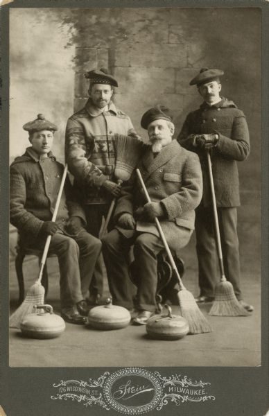 Full shot of four men posing with their curling equipment in sepia tone. Two sit on chairs, including John Johnston, interspersed with two others that stand. They all hold brooms in their warm clothes and hats. Three curling stones are placed on the floor close to their feet.