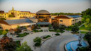 Bird's eye view of Alverno College facade and its front yard. Large letters on the awning say "Sister Joel Read Center." The yard features neatly-maintained landscaping and a blue-colored semi-circle fountain with a contemporary statue in its center. Sunlight bathes the rooftops.