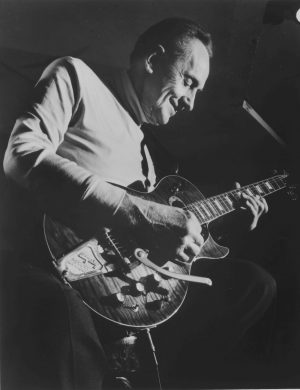 Grayscale low-angle shot of Les Paul in a sweater and trousers smiling as he plays guitar. His body faces to the right. His eyes gaze down at the guitar that is placed on his lap.